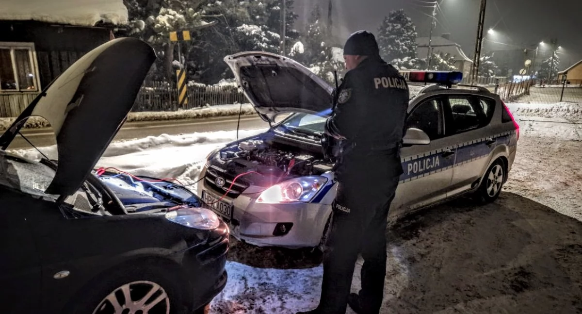Policjant na tle samochodów połączonych kablami rozruchowymi. Nie wiemy czy prawidłowo, wiemy, że się udało. Brawo policja.